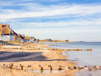 Sandstrand am Heiligenhafen Graswarder mit Holzpfosten, ruhiger Ostsee und traditionellen Reetdachhäusern entlang der Küste.