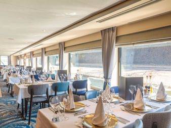 Eleganter Speisesaal an Bord der MS Lisabelle mit weiß gedeckten Tischen, blau gepolsterten Stühlen und großen Fenstern mit Blick aufs Wasser.
