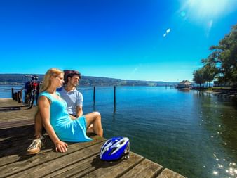 Two cyclists sitting on a wooden pier at Lake Constance with bicycles behind them. Blue helmet in foreground, calm lake and sunny sky visible.