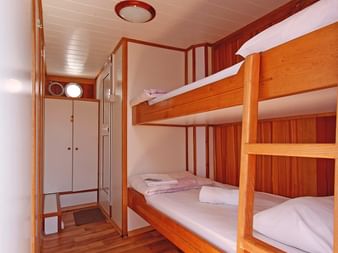 Kompakte Kabine auf der MS Mirabela mit Holz-Etagenbetten, weißer Bettwäsche, Schrank, Spiegel und Bullaugen. Holzverkleidung und Boden.