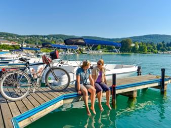 Zwei Radfahrer sitzen auf einem Holzsteg im Hafen von Velden mit den Füßen im türkisfarbenen Wasser. Ein Fahrrad steht daneben, Boote liegen dahinter.