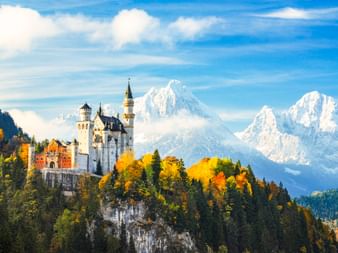 Schloss Neuschwanstein auf einem bewaldeten Hügel mit Herbstlaub, schneebedeckte Alpen im Hintergrund unter blauem Himmel mit weißen Wolken.