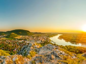 Panoramablick auf die Donau bei Hainburg bei Sonnenuntergang, mit felsigen Hügeln im Vordergrund und der Stadt im Tal unter goldenem Himmel.
