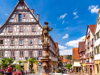 Marktplatz in Besigheim mit verziertem Brunnendenkmal, traditionellen Fachwerkhäusern und Besuchern, die den sonnigen Platz unter blauem Himmel genießen.
