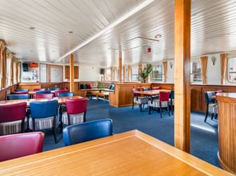 Geräumiges Salon-Restaurant an Bord der Floor van Aemstel mit Holzvertäfelung, blauem Teppich, roten und blauen Stühlen unter weißer Decke.