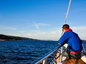 Person in blauer Jacke sitzt am Bug der MS Dalmatino und blickt auf das tiefblaue Adriatische Meer mit bewaldeter Küstenlinie in der Ferne.