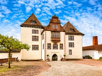 Schloss Fürstenberg mit weißen Mauern und braunen Verzierungen, mit zentralem Turm mit Uhr und Torbogen. Kopfsteinpflasterhof unter blauem Himmel.