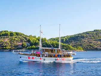 Motorschiff MS Dalmatino mit roten Fendern auf ruhigem blauen Wasser. Grüne terrassierte Hügel mit vereinzelten Gebäuden im Hintergrund.