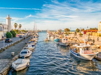 Kanal in Viareggio mit Fischerbooten an Steinmauern. Ein weißer Leuchtturm steht links, Palmen säumen das Ufer unter blauem Himmel.