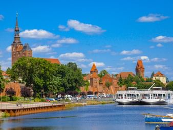 Blick auf Tangermündes mittelalterliche Silhouette mit Kirchturm und Backsteintoren an der Elbe, mit weißem Passagierschiff und blauem Himmel.