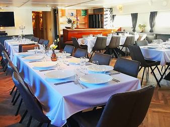 Elegantes Restaurant an Bord der MS Magnifique IV mit weiß gedeckten Tischen, dunklen Stühlen, Fischgrätparkett und Fenstern an den Wänden.