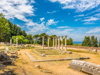 Antike griechische Ruinen des Asklepieion auf Kos mit stehenden Säulen, Steinfundamenten und verstreuten Ruinen unter blauem Himmel mit Meerblick.