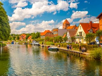 Kanal in Plau am See mit bunten Häusern, orangefarbenen Ziegeldächern, Kirchturm, vertäuten Booten und Holzsteg unter blauem Himmel.
