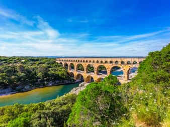 Pont du Gard, eine dreistöckige römische Aquäduktbrücke über den Gardon bei Remoulins, umgeben von üppiger grüner Vegetation unter blauem Himmel.