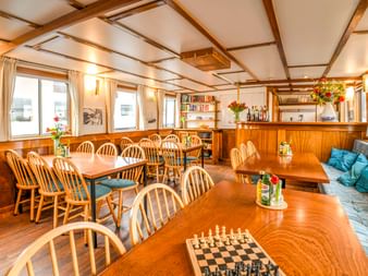 Heller Salon an Bord der MS Flora mit Holzdeckenbalken, Esstischen mit Windsor-Stühlen, Schachspiel, eingebauter Bar und blauer Polsterung.