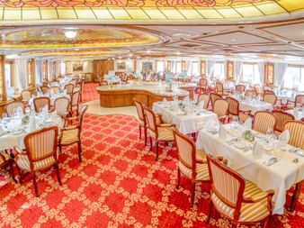 Elegantes Restaurant an Bord der MS Prinzessin Katharina mit weiß gedeckten Tischen, gestreiften Polsterstühlen, rotem Teppich und Decke.