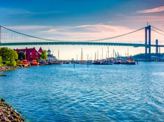 Älvsborg-Brücke über dem Hafen in Göteborg mit blauem Wasser, Booten und roten historischen Gebäuden am Klippan-Ufer unter Abendhimmel.