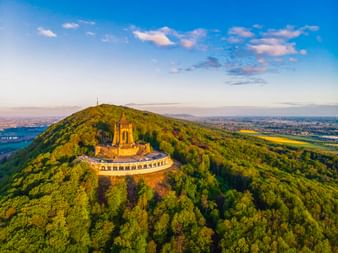 Luftaufnahme des Kaiser-Wilhelm-Denkmals auf einem bewaldeten Hügel in Porta Westfalica mit umliegender Landschaft und blauem Himmel.