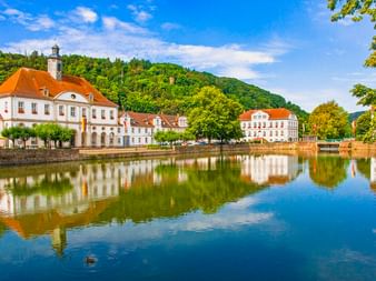 Weißes Barockrathaus mit orangem Dach in Bad Karlshafen spiegelt sich im ruhigen Wasser der Weser, umgeben von historischen Gebäuden und grünen Hügeln.
