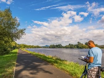 Radfahrer mit Fahrrad sitzt im Gras neben asphaltiertem Weg entlang der Elbe, lesend unter blauem Himmel mit weißen Wolken und grünen Bäumen.