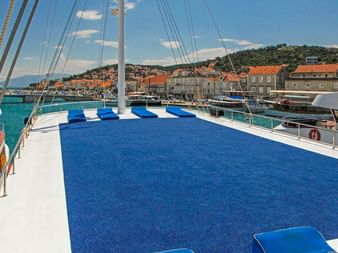 Sonnendeck der MS Azimut mit blauem Teppich und Kissen, Mast und Takelage sichtbar. Küstenstadt mit orangefarbenen Dächern im Hintergrund.