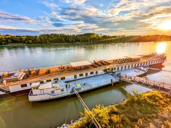 Flusskreuzfahrtschiff MS Prinzessin Katharina liegt bei Sonnenuntergang an der Donau. Das weiße Schiff mit Holzdeck hat Sonnenliegen und ist über eine Gangway mit dem Ufer verbunden.