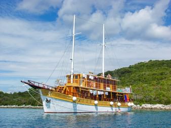 MS Planka, ein hölzernes Zweimaster-Segelschiff mit weißem Rumpf und gelb-braunem Aufbau, vor Anker in ruhigem blauen Wasser mit grünen Hügeln.