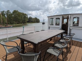 Esstisch mit Stühlen auf dem Sonnendeck des Schiffes Floor van Aemstel, mit Flussblick und Steuerhaus im Hintergrund unter bewölktem Himmel.