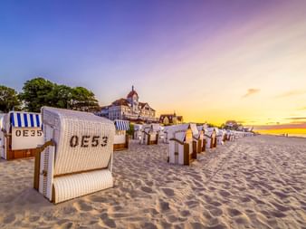 Reihen weißer Strandkörbe am Sandstrand von Kühlungsborn bei Sonnenuntergang. Historisches Gebäude mit Turm im Hintergrund unter buntem Himmel.