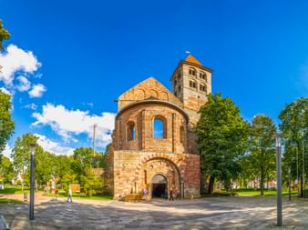 Romanische Kirchenruine mit runder Apsis und quadratischem Turm in Bad Hersfeld, umgeben von grünen Bäumen und gepflastertem Platz unter blauem Himmel.