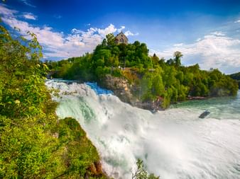 Rheinfall bei Schaffhausen mit herabstürzendem Wasser, üppiger grüner Vegetation und einem historischen Gebäude auf einem Felsvorsprung.