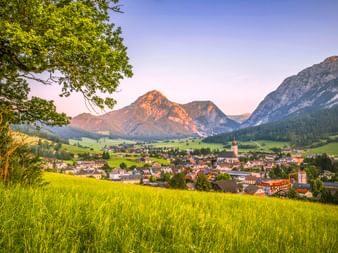 Alpendorf Gröbming in der Steiermark in grünem Tal mit Kirchturm, umgeben von Wiesen und dramatischen Berggipfeln unter Abendhimmel.