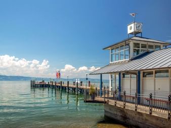 Hafengebäude in Kressbronn mit Uhrturm und Metalldach am Bodensee. Holzsteg mit Anlegepfosten führt ins ruhige Wasser.