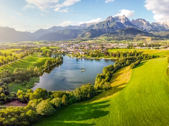 Aerial view of Saalfelden with green meadows, a lake, and snow-capped mountains in the background under a blue sky with clouds.