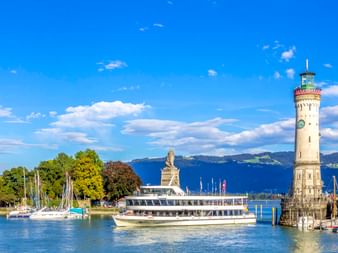 Lindauer Hafeneinfahrt mit weißem Leuchtturm und Löwenstatue. Ein Passagierschiff fährt zwischen ihnen auf dem Bodensee bei blauem Himmel.