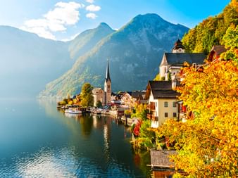 Hallstatt mit Kirchturm und traditionellen Häusern am Seeufer, umgeben von Herbstlaub und Bergen unter blauem Himmel.