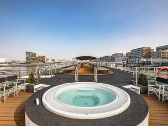 Sonnendeck der MS Swiss Crown mit rundem Whirlpool, Liegestühlen und Holzdeck. Städtische Uferpromenade mit modernen Gebäuden im Hintergrund.