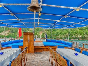 Sonnendeck der MS Thalassa mit Holzbänken, blauen Kissen und blauer Markise. Küstenlandschaft mit grünen Hügeln im Hintergrund.
