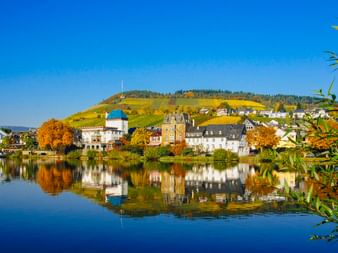 Stadt Traben-Trarbach an der Mosel mit Herbstlaub. Historische Gebäude spiegeln sich im ruhigen Wasser, dahinter Weinberge an den Hügeln.