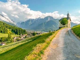 Kleine Kapelle mit Glockenturm auf grünem Hügel mit Blick auf Toblach in Südtirol. Schotterweg führt zur Kapelle mit Bergen im Hintergrund.