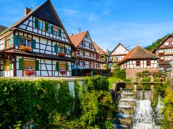 Traditionelle Fachwerkhäuser in Reichental mit bunten Fensterläden entlang eines kleinen Wasserfalls. Die historischen Gebäude haben weiße Wände mit dunklen Balken.