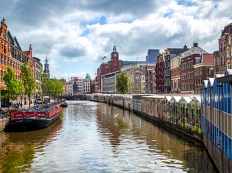 Amsterdamer Kanal gesäumt von bunten historischen Gebäuden und Kirchtürmen unter bewölktem Himmel. Ein Boot liegt links am Ufer.
