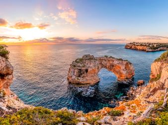 Es Pontàs natural stone arch in Mallorca at sunset. Rocky cliffs frame the arch formation rising from blue Mediterranean waters.