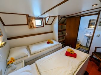 Zweibettkabine auf Floor van Aemstel mit zwei Einzelbetten, weißer Bettwäsche, roten Handtüchern, gelben Hüten, Holzverkleidung, Bullauge und eigenem Bad.