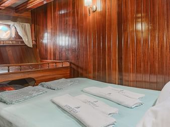 Doppelkabine auf der MS Thalassa mit holzgetäfelten Wänden, Bett mit hellgrüner Bettwäsche, weißen Kissen, Bullaugen und Einbauschränken.