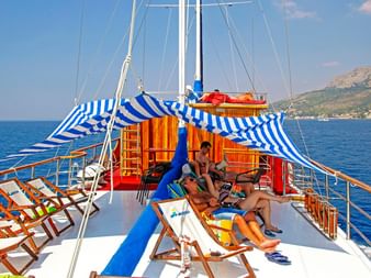 Sonnendeck der MS Mirabela mit Gästen auf Liegestühlen. Blau-weiß gestreifte Markisen spenden Schatten, blaues Meer und Küstenhügel im Hintergrund.