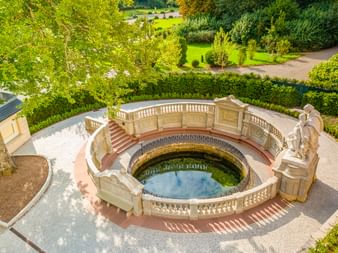 Luftaufnahme der Donauquelle, ein kreisförmiger Steinbrunnen mit kunstvollen Balustraden und Skulpturen, umgeben von grüner Parklandschaft.