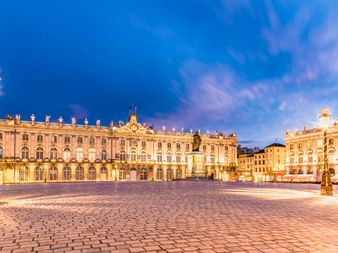 Place Stanislas in Nancy bei Abenddämmerung mit beleuchteten historischen Gebäuden, verzierten Straßenlaternen und Kopfsteinpflaster unter blauem Himmel.