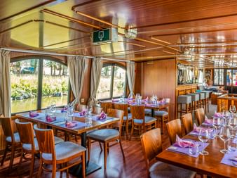 Innenraum des MS L'Estello Restaurants mit Holztischen, lila Servietten und Weingläsern. Große Fenster mit Vorhängen bieten Kanalblick.