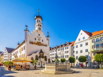 Historisches weißes Kemptener Rathaus mit Uhrenturm und verzierter Fassade auf Kopfsteinpflasterplatz. Brunnen und Straßencafé unter blauem Himmel.